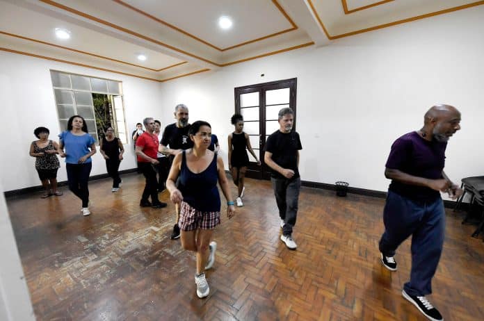 Grupo de alunos pratica passos de forró e samba rock durante aulas de dança gratuitas em Guarulhos, em um amplo salão iluminado com instrutores ao centro.