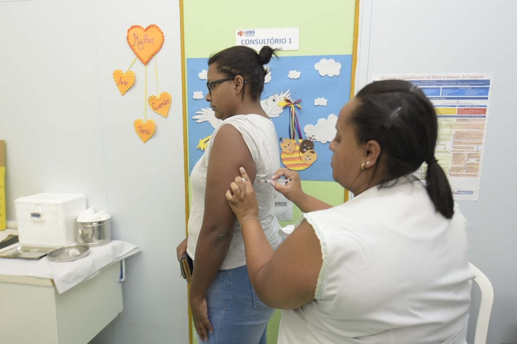 Profissional de saúde aplicando vacina no braço de uma pessoa em uma UBS de Guarulhos. Ao fundo, frascos de vacina e materiais de proteção, simbolizando a campanha de vacinação contra sarampo e febre amarela.