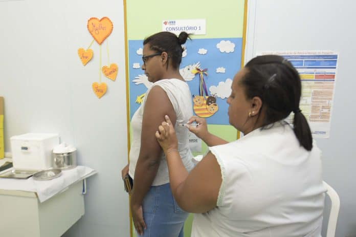 Profissional de saúde aplicando vacina no braço de uma pessoa em uma UBS de Guarulhos. Ao fundo, frascos de vacina e materiais de proteção, simbolizando a campanha de vacinação contra sarampo e febre amarela.