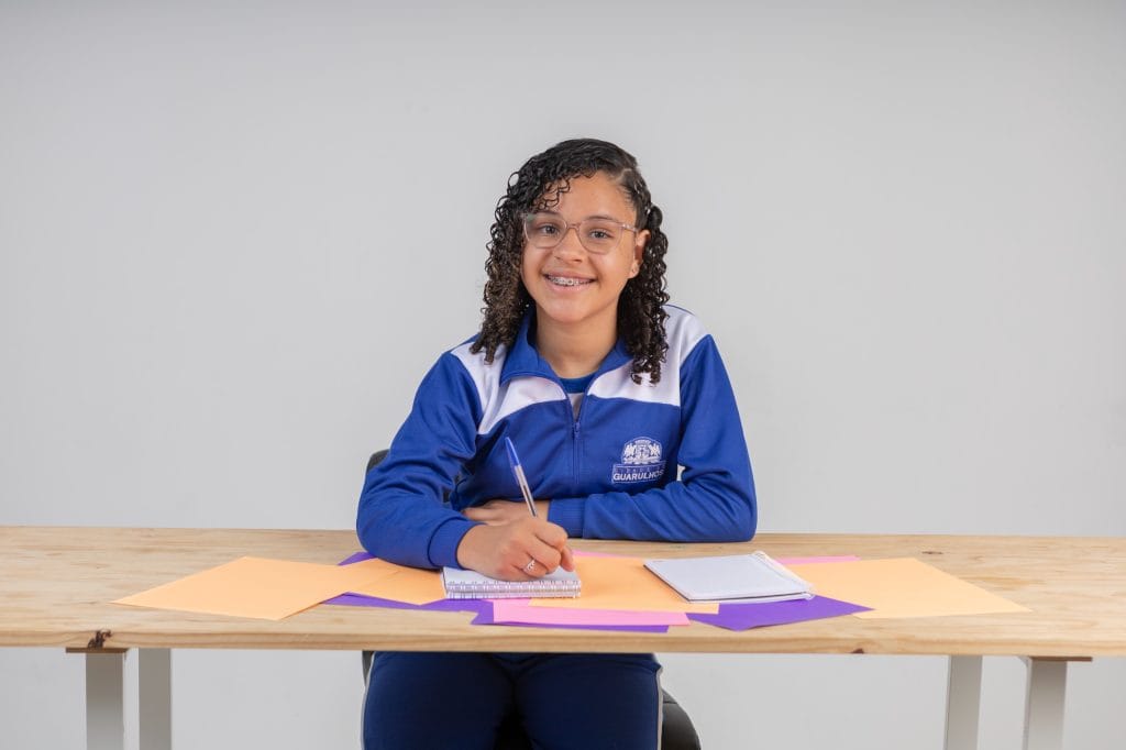 Estudantes da rede municipal de Guarulhos sorridentes, utilizando o uniforme escolar azul e carregando mochilas, simbolizando o retorno às aulas nas escolas da prefeitura.