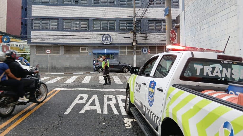 Agente de trânsito de Guarulhos, vestindo uniforme oficial, sinaliza a travessia de estudantes em uma faixa de pedestres em frente a uma escola municipal. Ao fundo, o fluxo de veículos organizado e cones de sinalização delimitando a área de embarque e desembarque.